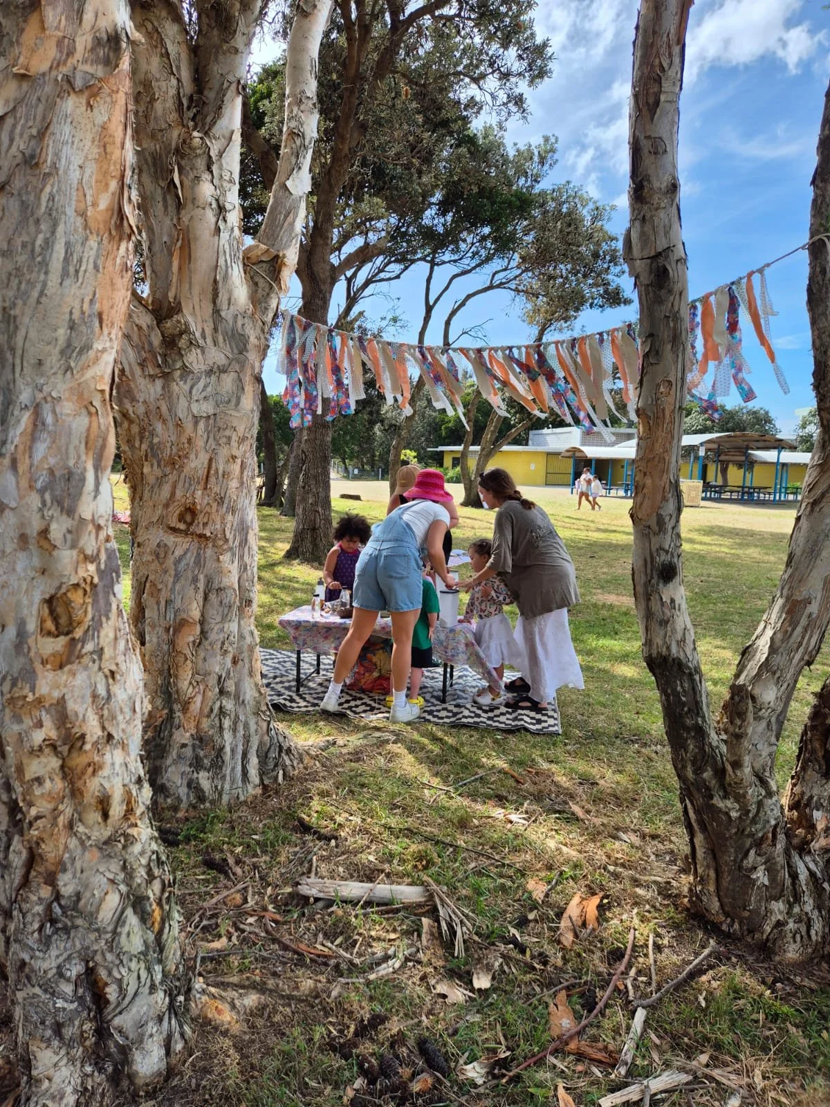 People gathering at a picnic table under trees in a park, with colorful fabric banners hanging between the trees and a playground in the background.