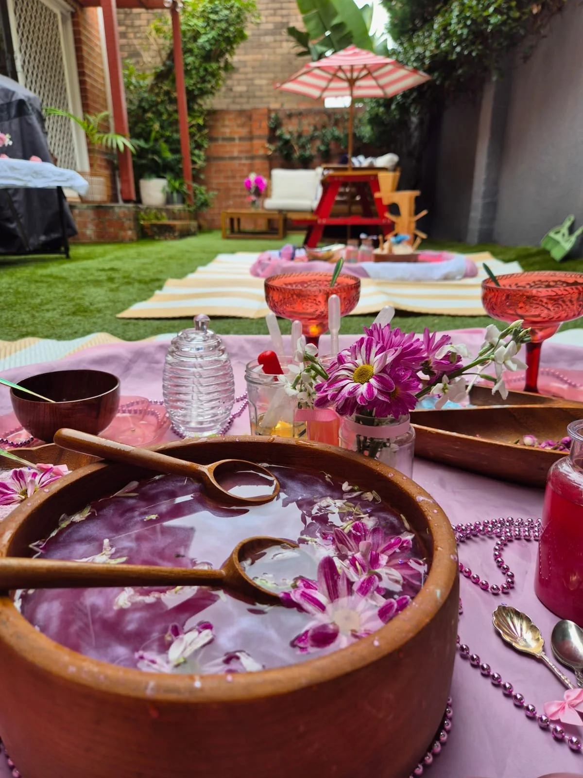 Outdoor summer party setup with a pink tablecloth, flower decorations, bowls, glasses, and a large wooden bowl with a floral drink. In the background, there are striped blankets, chairs, potted plants, and a striped umbrella on a grassy backyard.