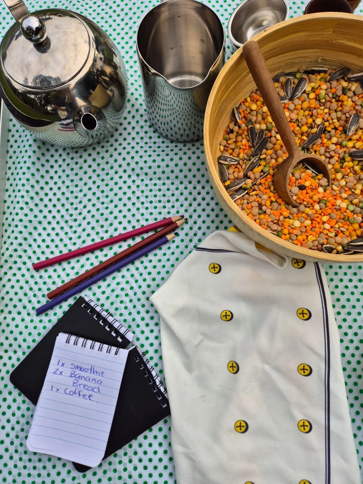 Table with various kitchen items, including a teapot, a metal pitcher, and a bowl of mixed beans and seeds with a wooden scoop, on a green polka-dot cloth. There are colored pencils, a notepad with a handwritten shopping list, and a white cloth with 