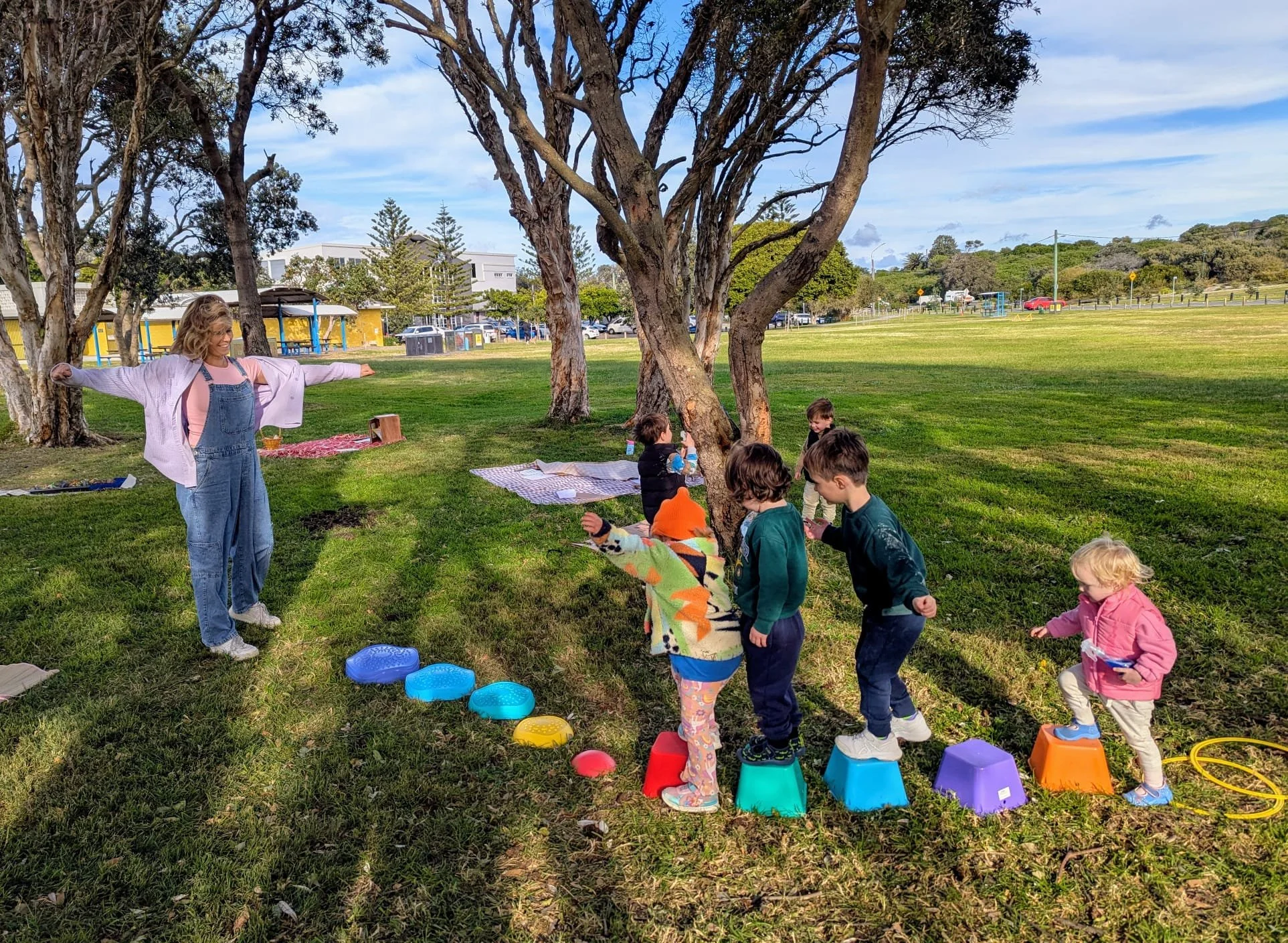 Children participating in an outdoor activity in a park with a woman supervising. The children are standing on colorful stepping stones arranged in a line under trees, with some holding hands. The background shows grassy fields, trees, and distant buildings.