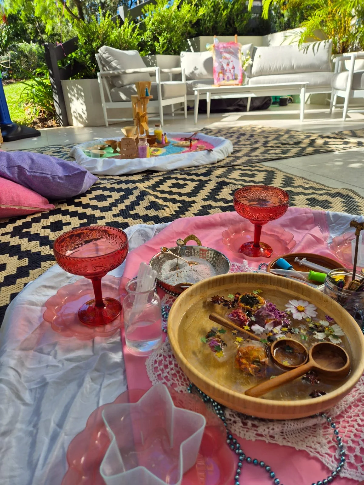 Outdoor table setting with pink glasses, flowers, and decorative items on a tablecloth, with a background of outdoor furniture and greenery.