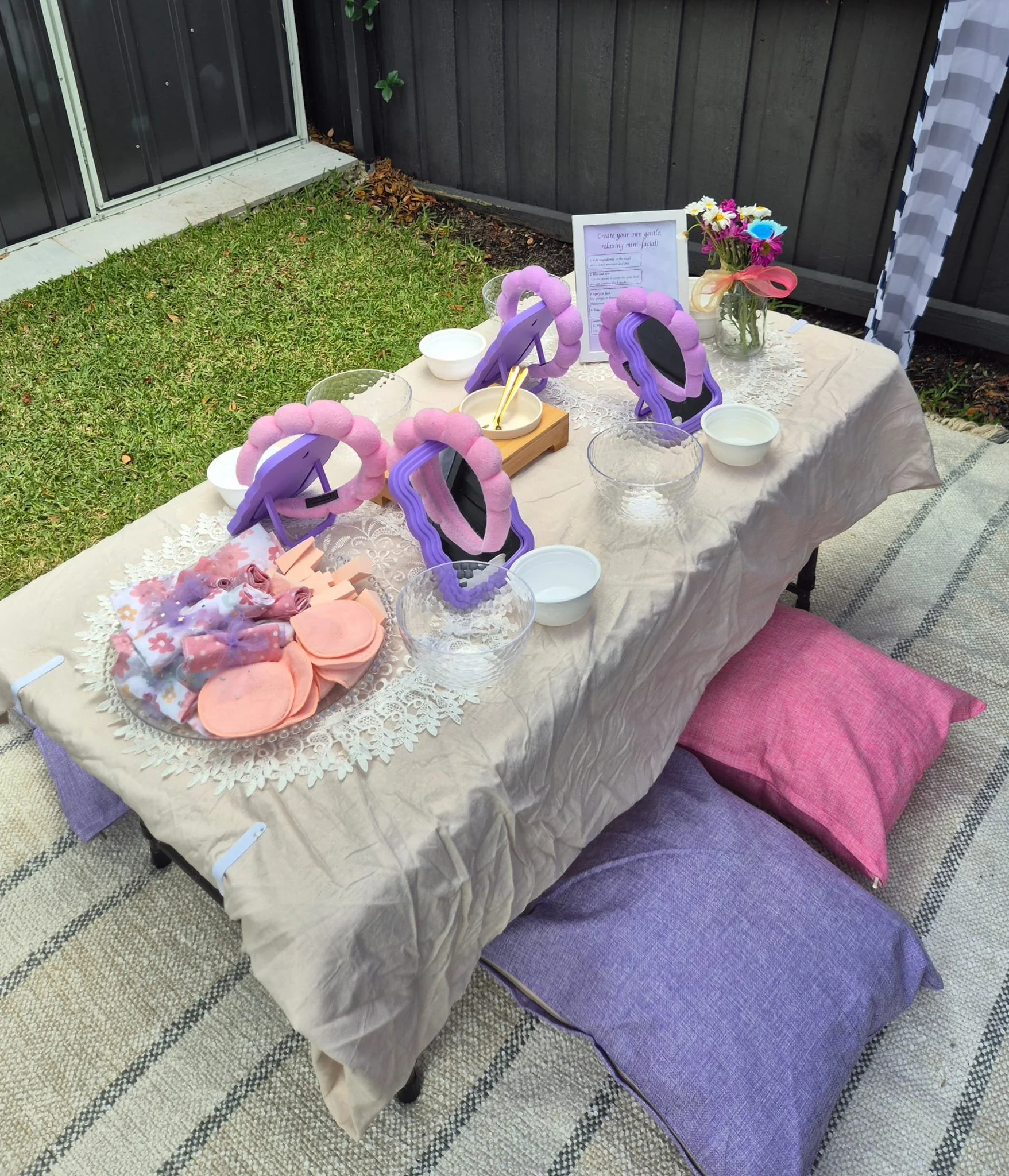 A table set outdoors with a beige tablecloth, featuring mini mirrors decorated with pink and purple felt, small bowls, a container with a flower arrangement, and a sign providing instructions for creating a mini facial. The background shows a grassy area and a black wooden fence.