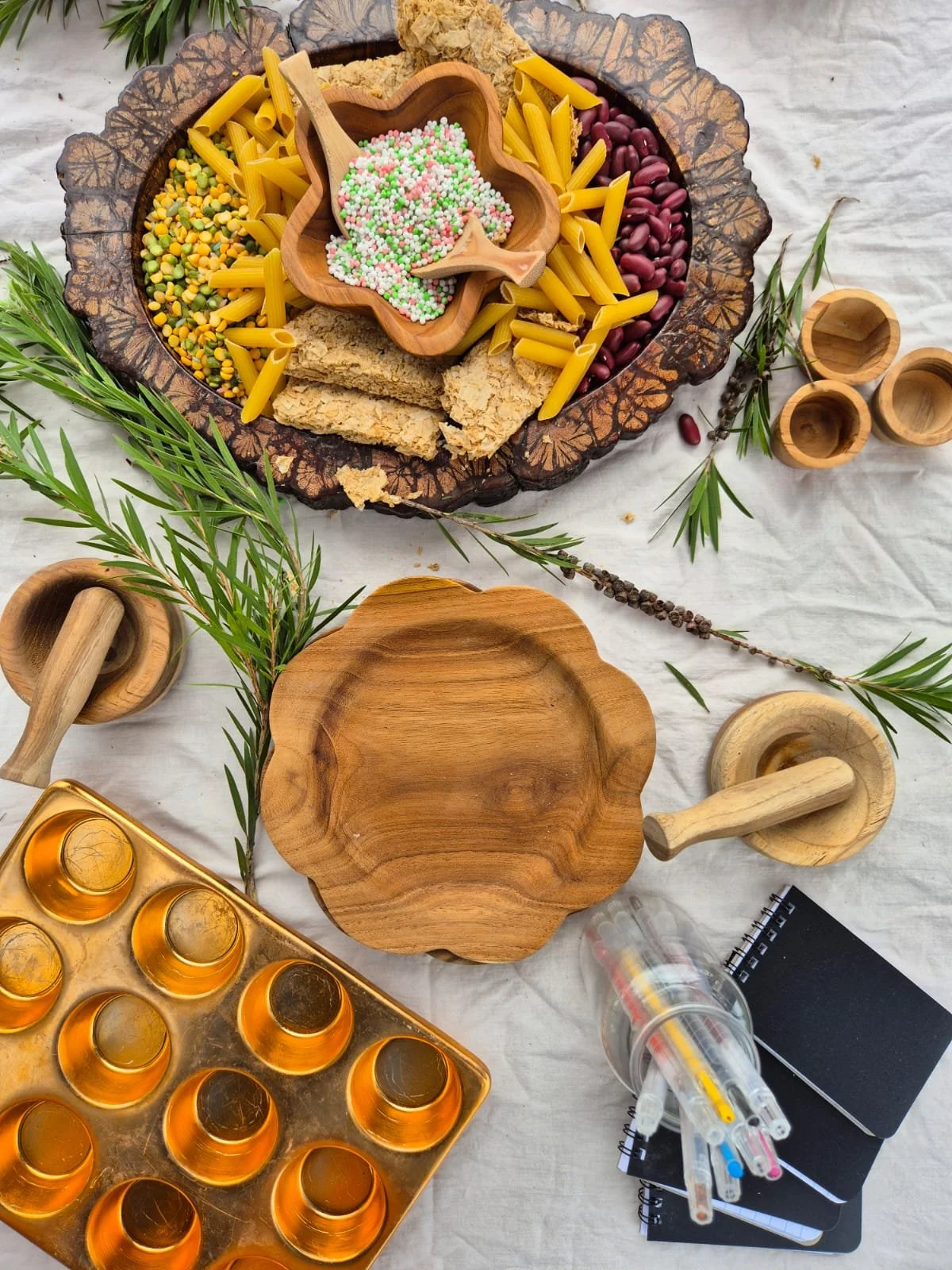 A wooden platter with colorful beans, pasta, crushed crackers, and sprinkles, surrounded by small wooden bowls, sprigs of greenery, a notebook, syringes, and a plastic container on a white surface.