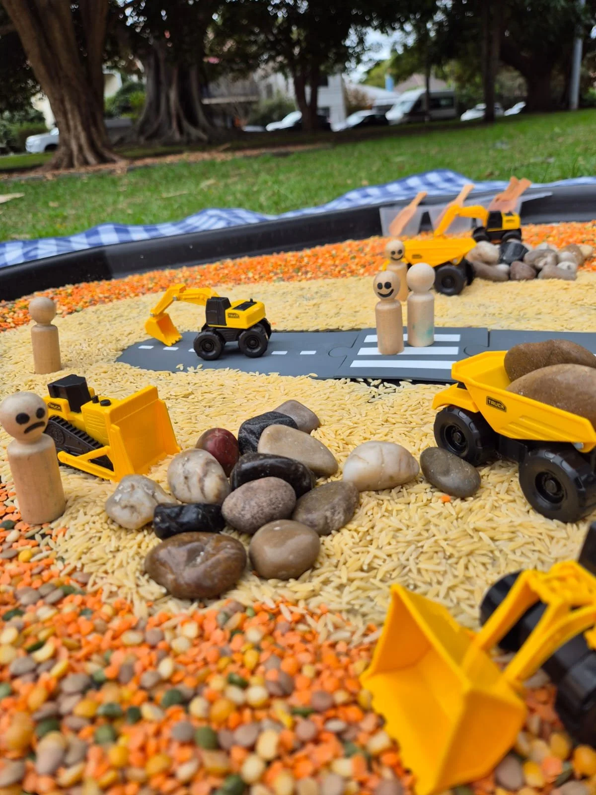 A miniature construction site with toy yellow dump trucks, construction figures, rocks, and grains of rice in an outdoor setting with trees in the background.