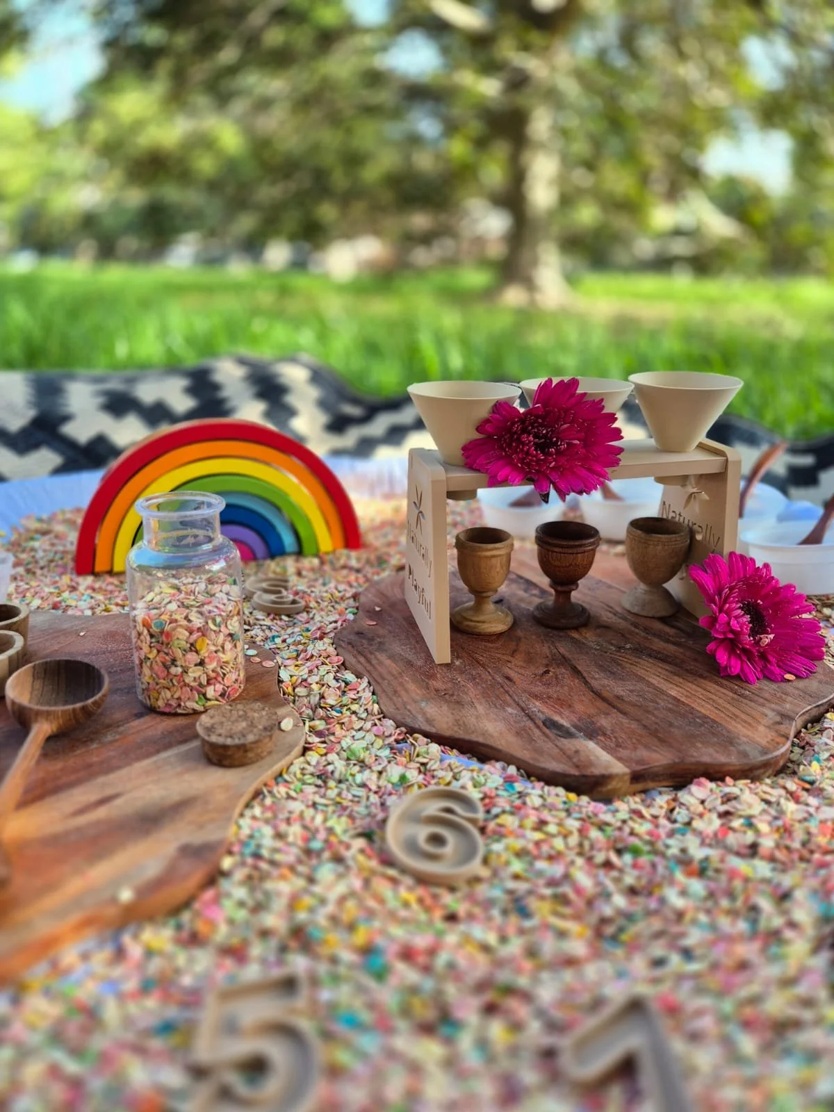 A colorful outdoor scene featuring a wooden table laden with rainbow-themed decorations, pink flowers, and assorted wooden cups and bowls. A rainbow display, jar of colorful sprinkles, and various decorative items are visible, set against a lush green backdrop with trees.