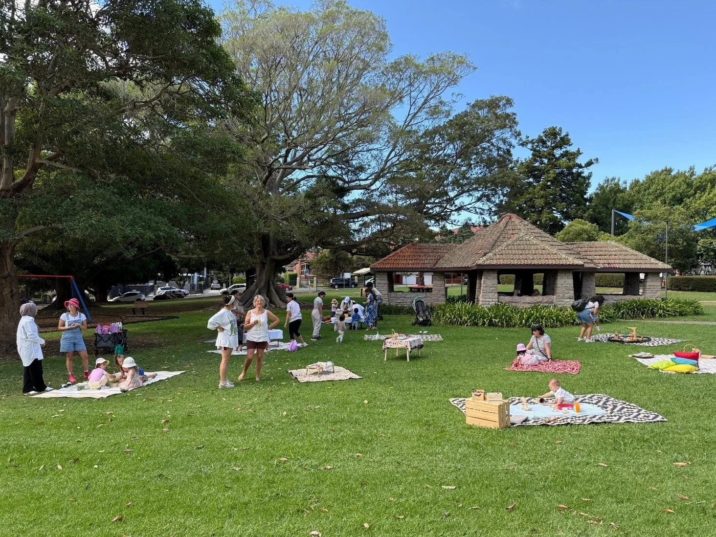 A park scene with a group of people, including children and adults, gathered on the grass around picnic blankets and tables. Some children are playing, and adults are chatting. There are large trees providing shade, and a small pavilion with a tiled roof is in the background. The sky is blue with no clouds.