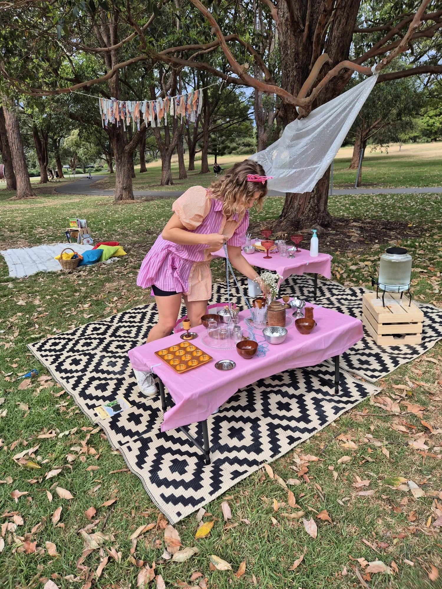 A woman setting up a small outdoor market or craft fair in a park. The table is covered with a pink tablecloth and displays various small bowls, glassware, and decorative items. A black and white patterned rug is underneath the table. Behind her, the