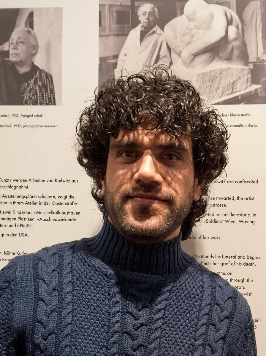 Rafi Rasooli, a portrait artist, A man with curly dark hair and a beard taking a selfie in front of a museum display with black-and-white photos and text about art and artists.