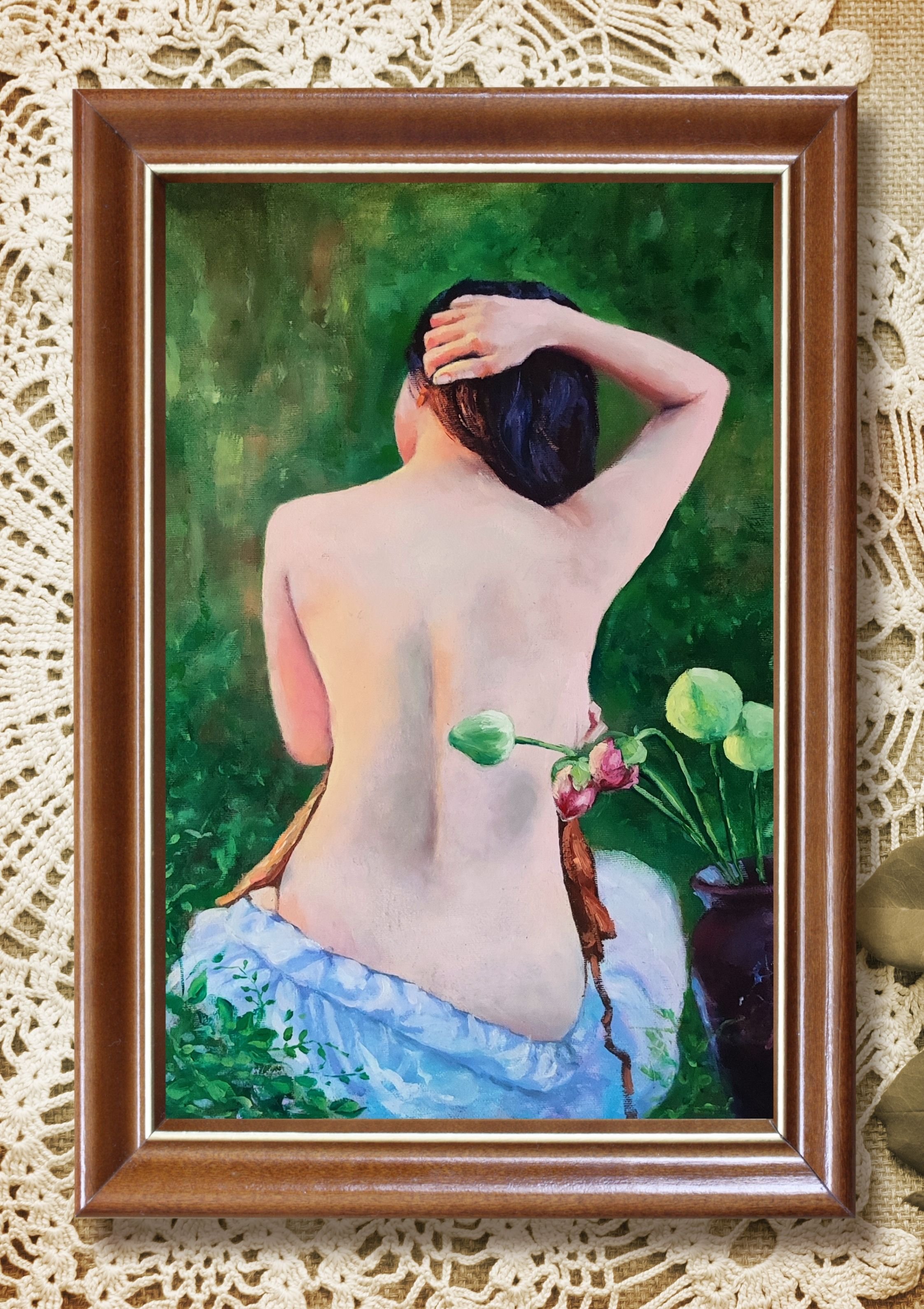 A painting of a nude woman seen from behind, with her left hand on her head, dark hair, and a greenery background, framed in wood, placed on a lace tablecloth.