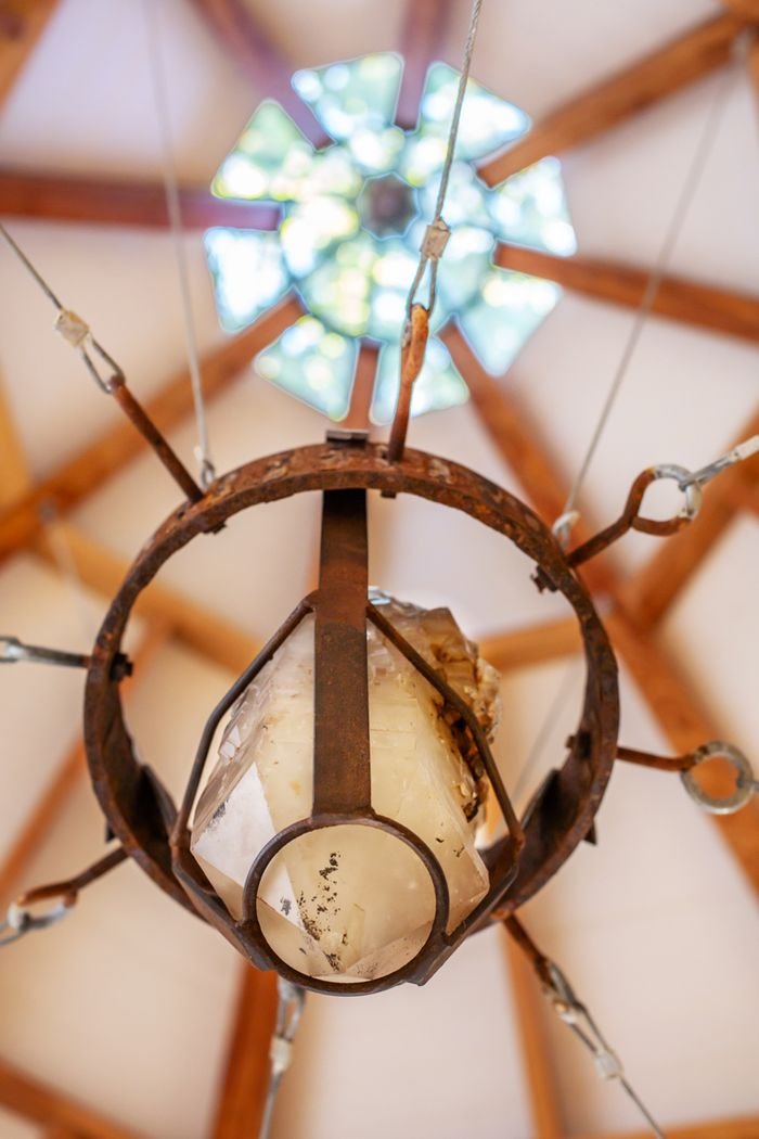 Close-up of an old, rusty chandelier with a frosted glass cover, hanging from a wooden ceiling with exposed beams, view from below with a circular window and blue sky in the background.