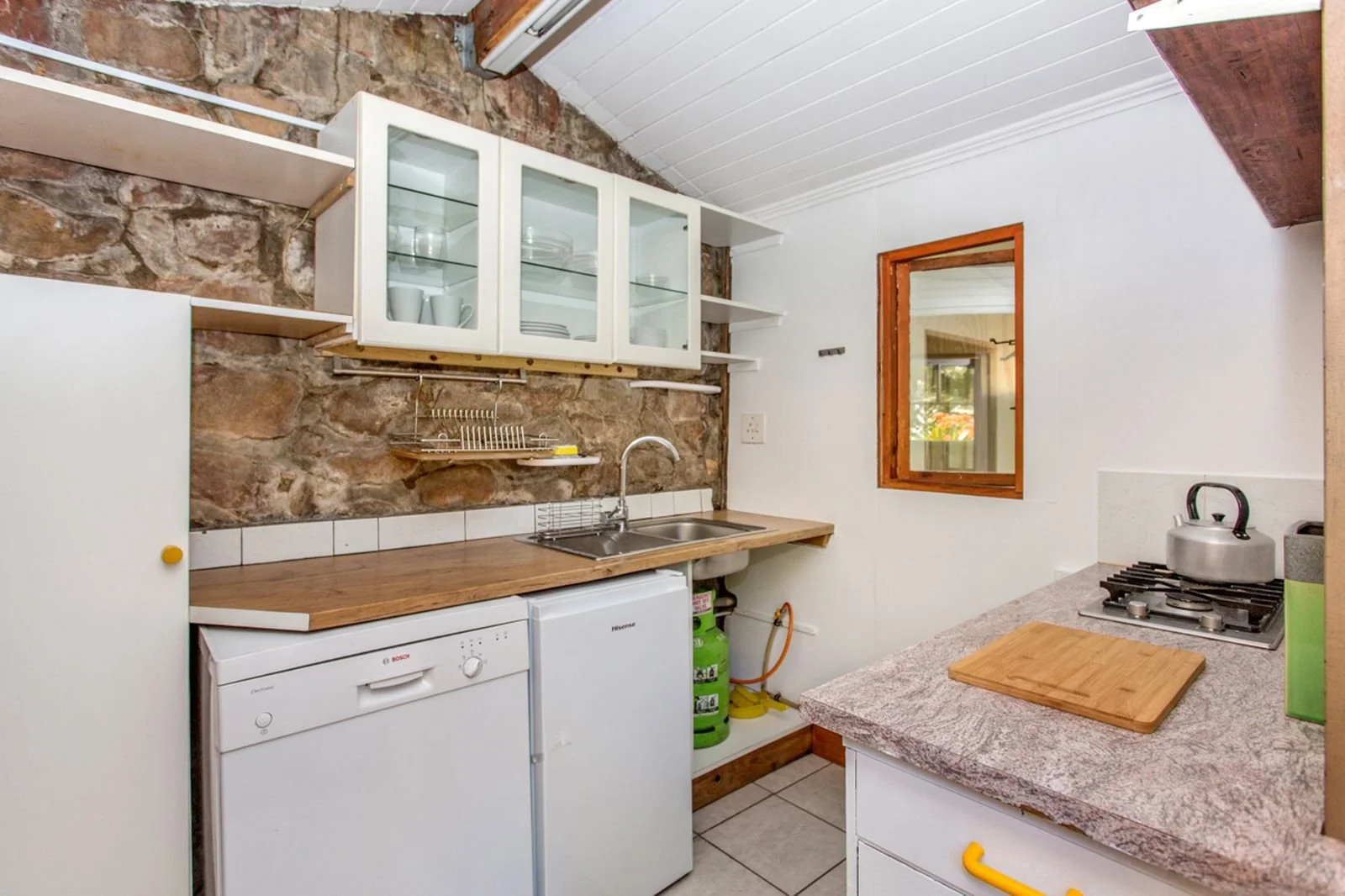 A small kitchen with a stone wall, white cabinetry, a dish rack, a double sink, a washing machine, gas stove, and wooden countertops.