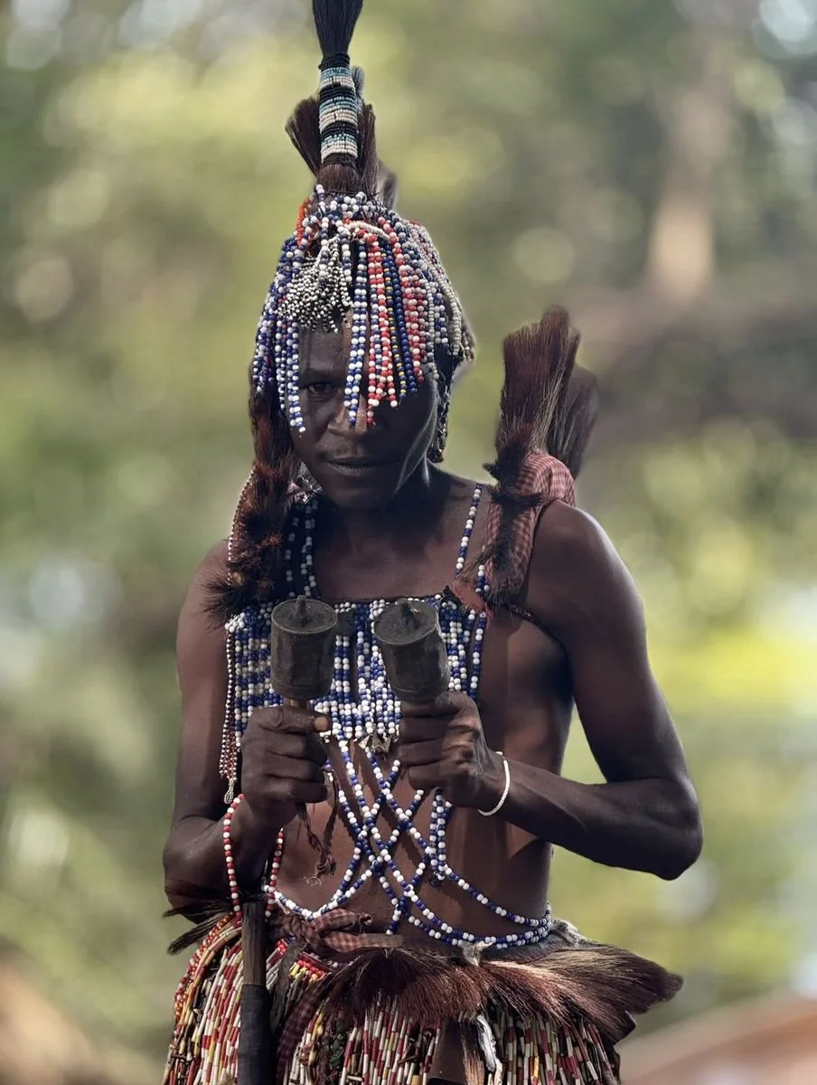 A man in traditional African attire, wearing beaded jewelry and a beaded headdress, holding two wooden objects with a forest background.
