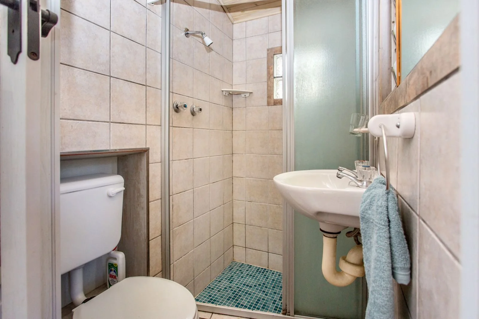 Small bathroom with a separate shower stall, a wall-mounted sink with a towel hanging from it, and a toilet with the tank visible through a built-in shelf. The walls are tiled, and there is a small frosted window inside the shower.