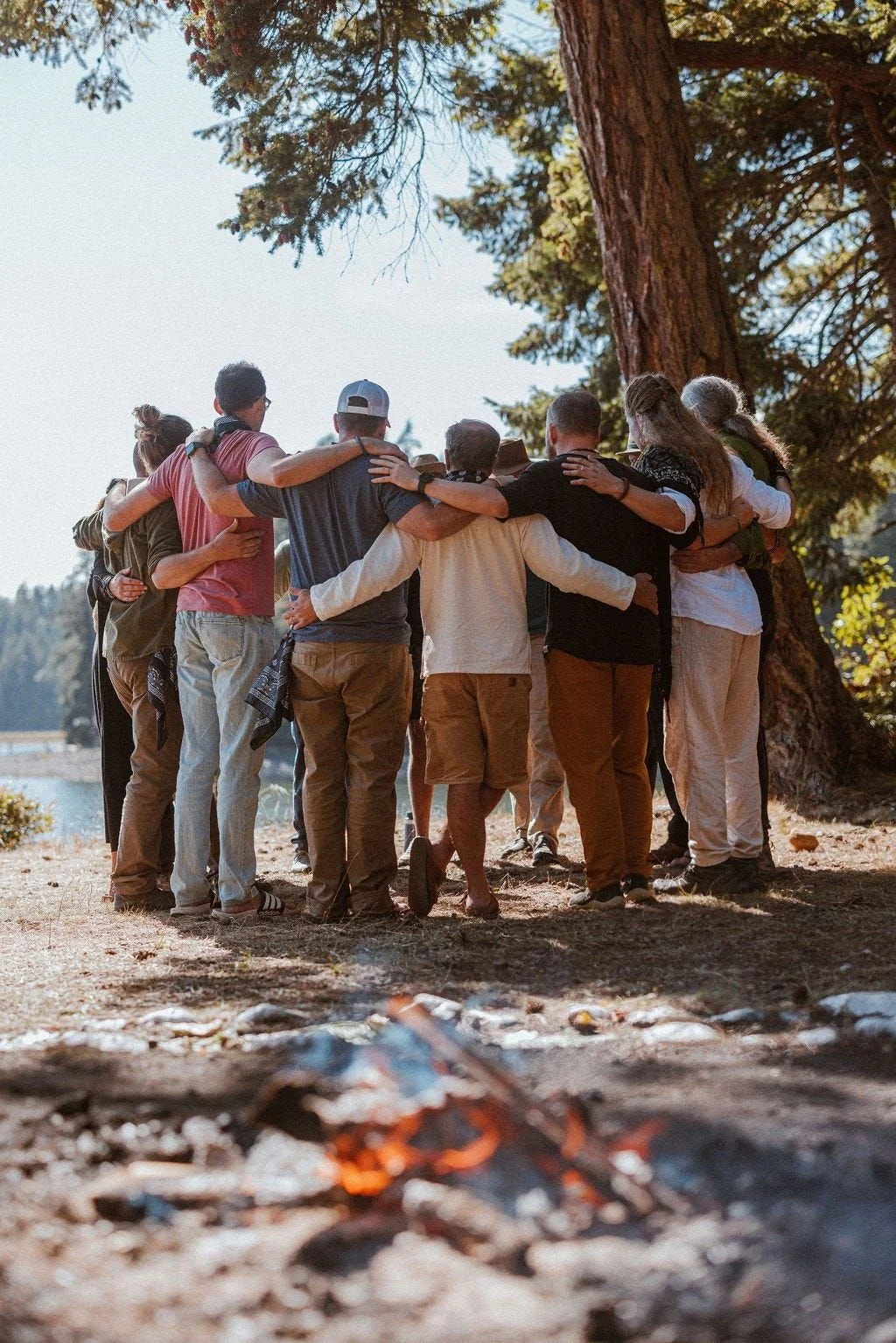 Group of people hugging each other in a circle outdoors near a large tree, with a campfire in the foreground and a scenic background of trees and possibly a lake.