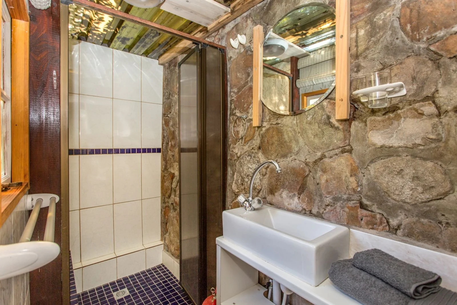 Bathroom with stone wall, small sink, shower with glass door, and window with wooden frame.