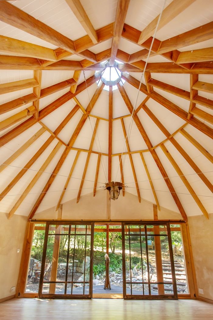 Interior view of a circular wooden ceiling with a central skylight, wooden beams radiating outward, and sliding glass doors looking onto a garden outside.