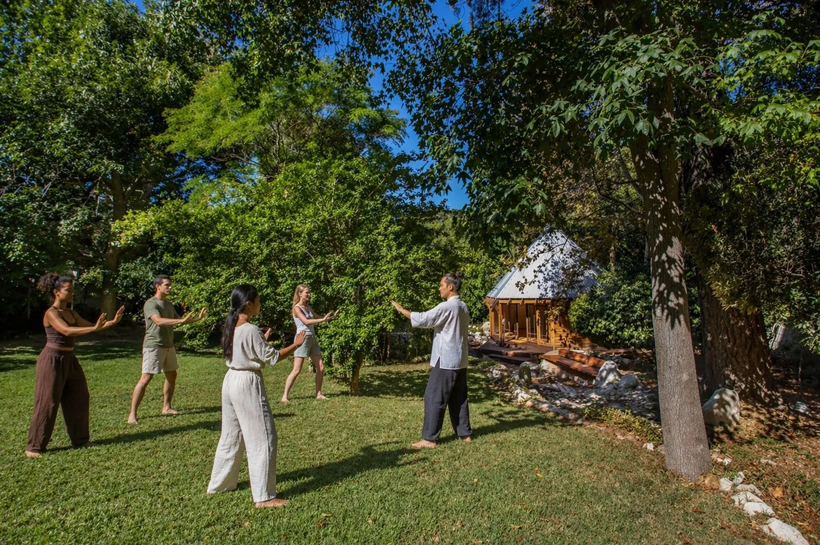 A group of five people at Akasha practicing tai chi or similar martial arts, led by an instructor in front of a healing temple, with mature trees and blue sky in the background.