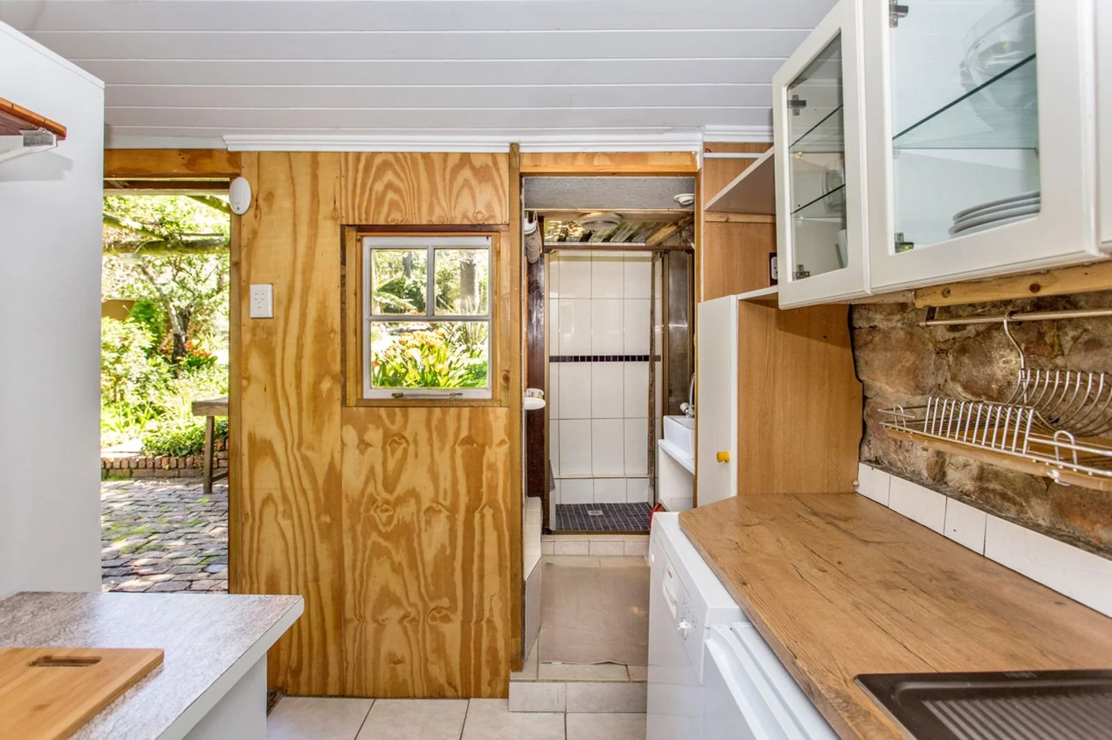 Kitchen with wooden cabinetry and countertops, a small window overlooking a garden, and a view into a bathroom with a shower.