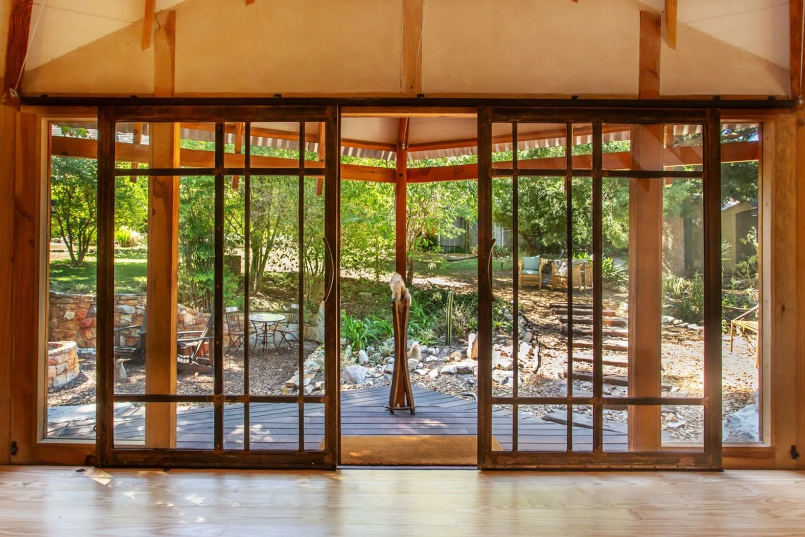 View of a backyard through wooden-framed glass sliding doors with a screen, showing outdoor furniture, a garden with trees, rocks, and a wooden deck.