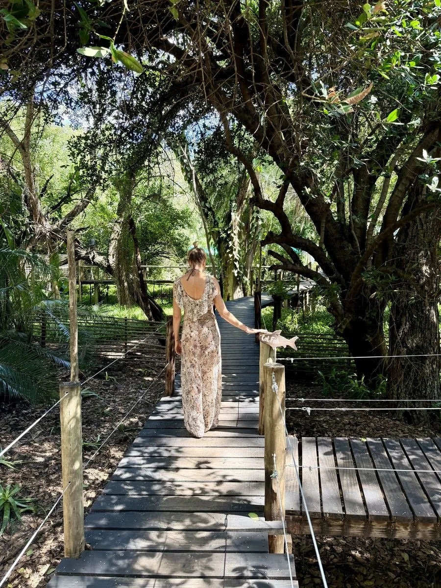 A woman in a beige and brown floral dress is walking on a wooden boardwalk through a lush green forest, with large trees and dense foliage on either side.