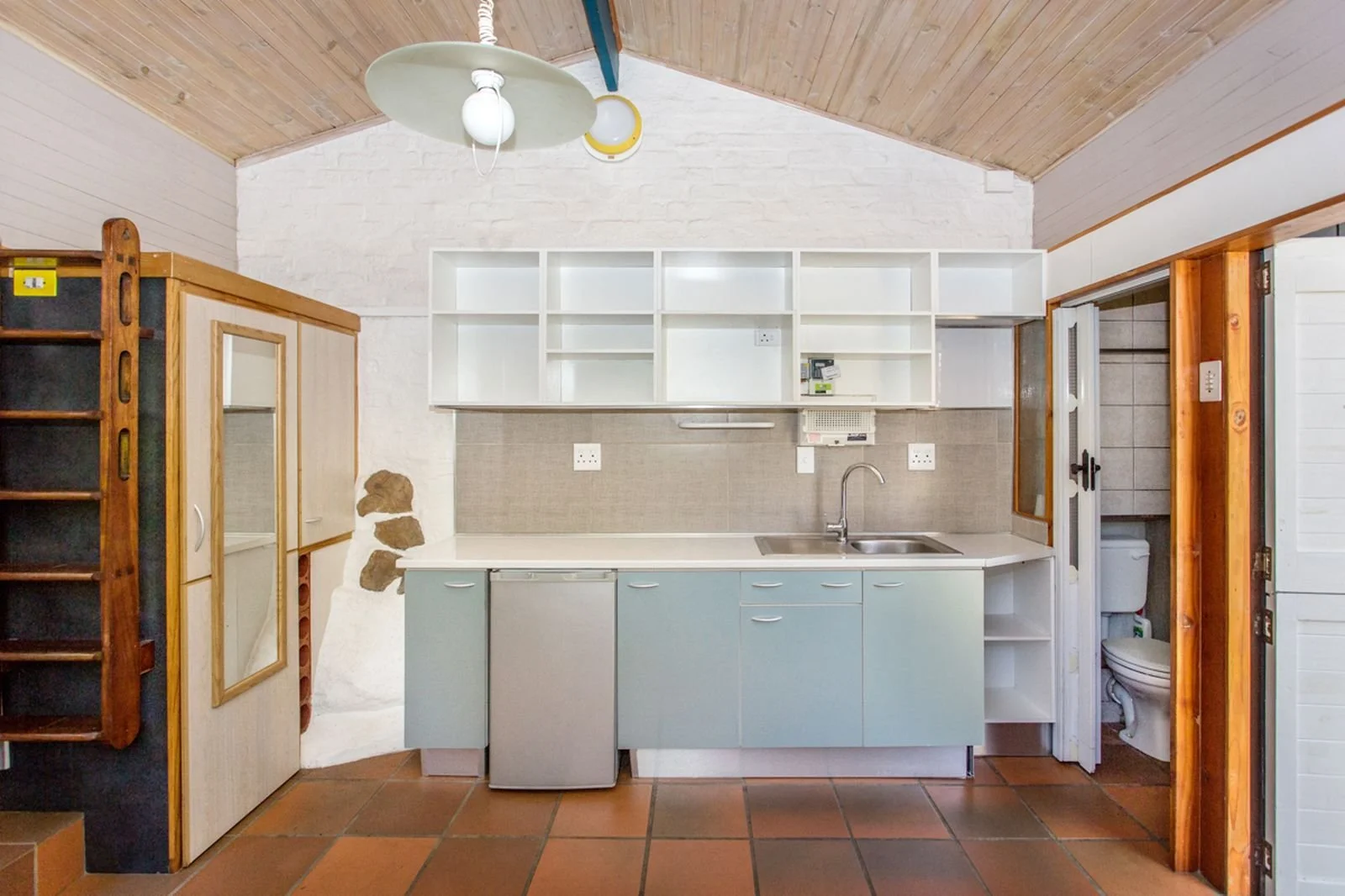 Small kitchen with white upper cabinets, light blue lower cabinets, a sink, and a beige backsplash. To the right, there's a bathroom with a toilet visible through an open door. The ceiling is wooden, and the floor has brown tiles. There is a white and green light fixture hanging from the ceiling.