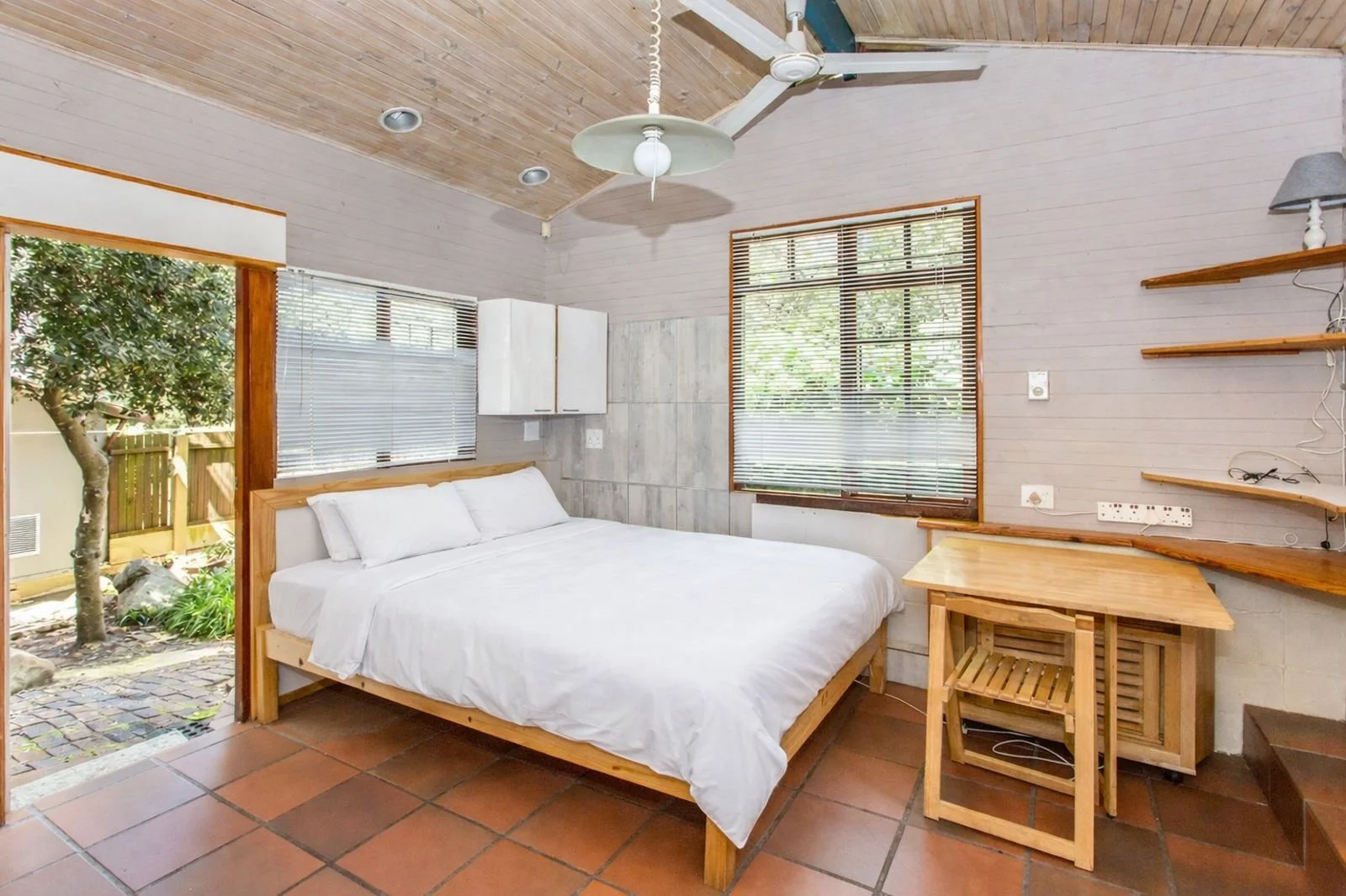 A cozy bedroom with a wooden bed and white bedding, large windows with blinds, a small wooden desk and stool, floating shelves, a ceiling fan, and view of a yard with a tree outside.