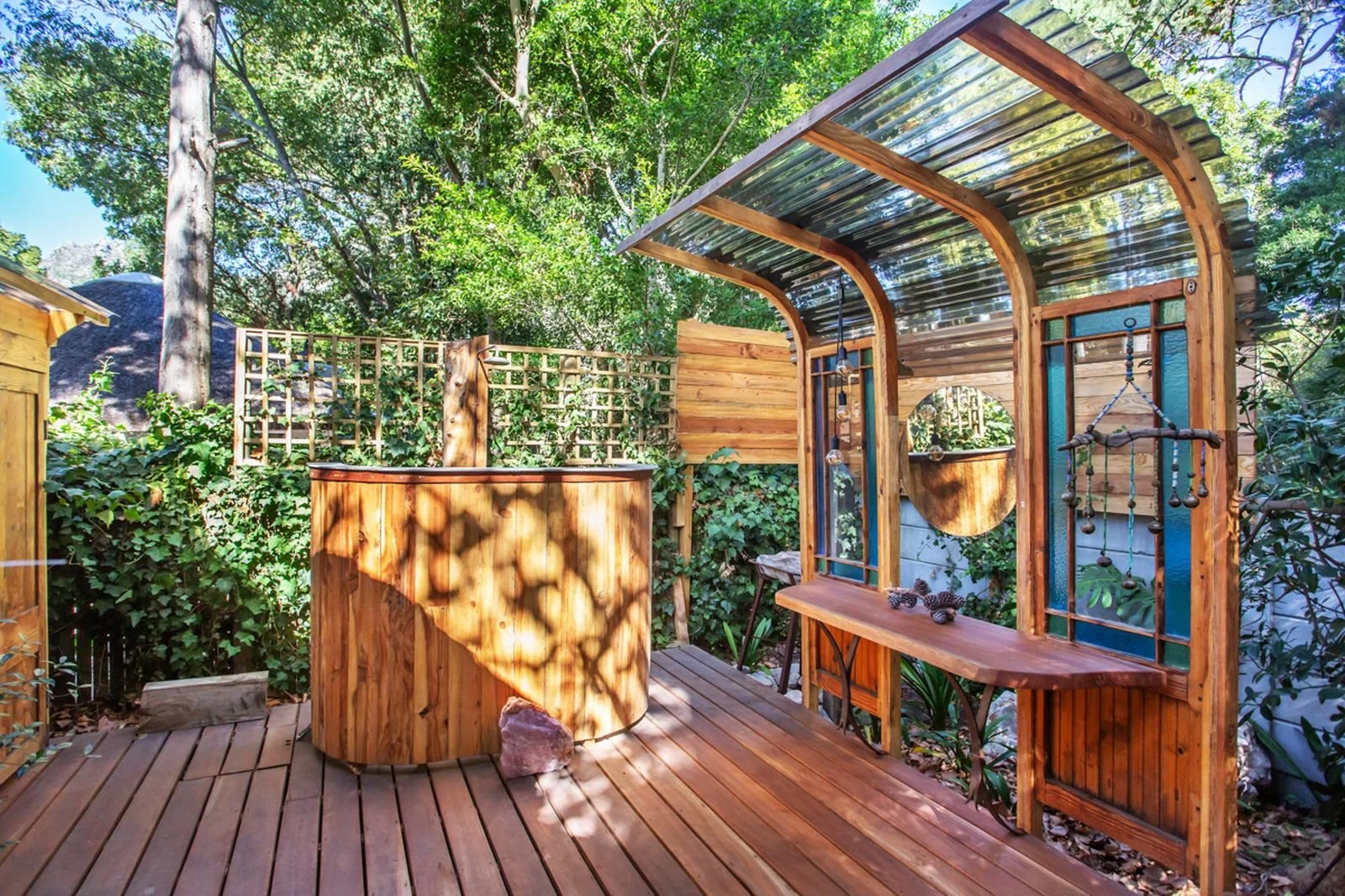 A backyard wooden deck with a rustic, outdoor shower and privacy fencing, surrounded by lush green trees and plants.