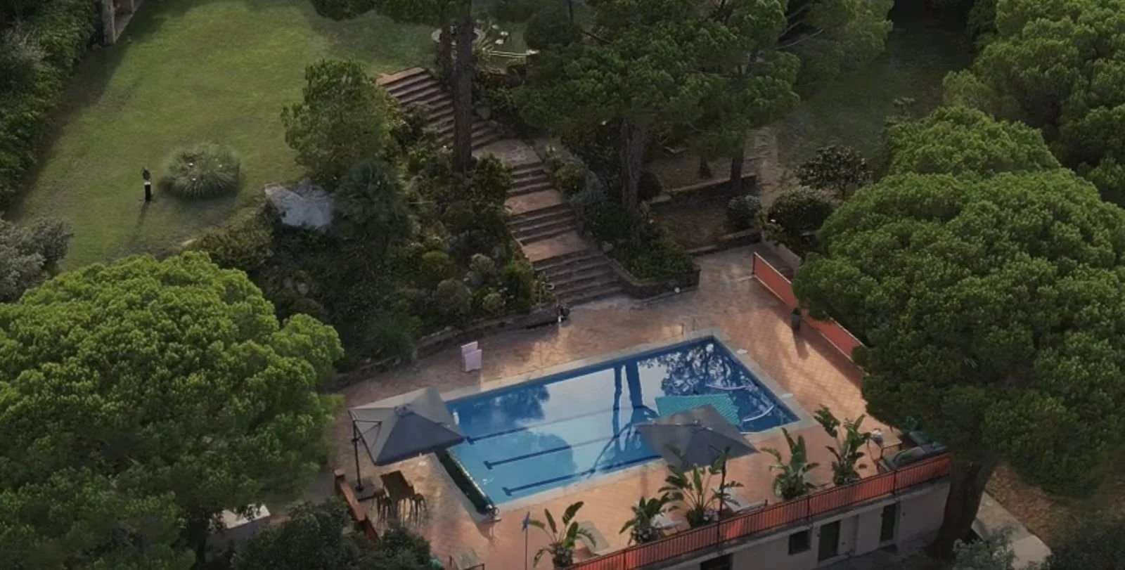 Aerial view of a backyard featuring a swimming pool surrounded by a patio with outdoor furniture, lush green trees, and a garden area with steps leading up a hill.