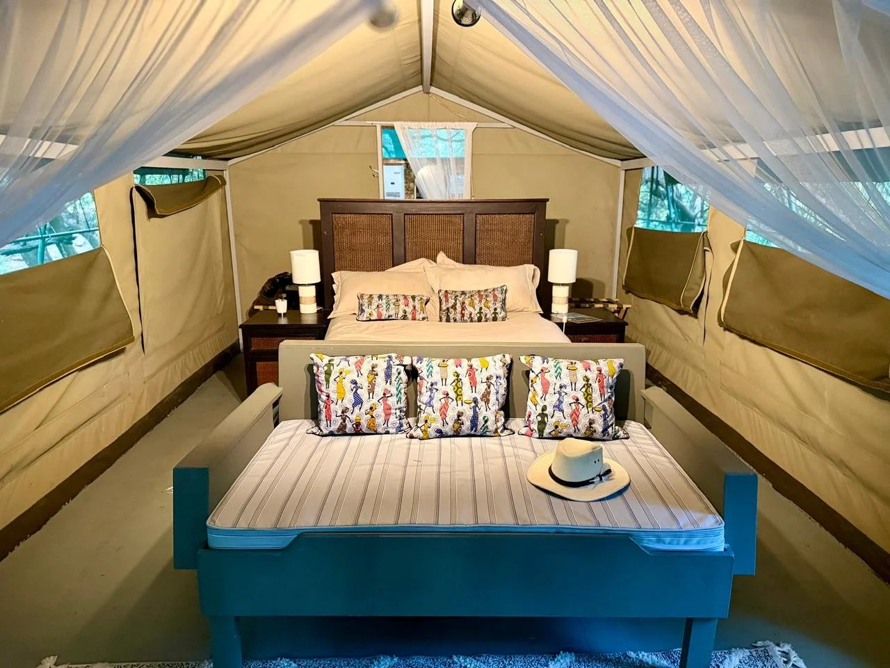 Inside a luxurious safari tent, a bed with decorative pillows, bedside tables with lamps, and a separate daybed with pillows and a white hat. The tent has beige walls, a high ceiling, and windows with green curtains.