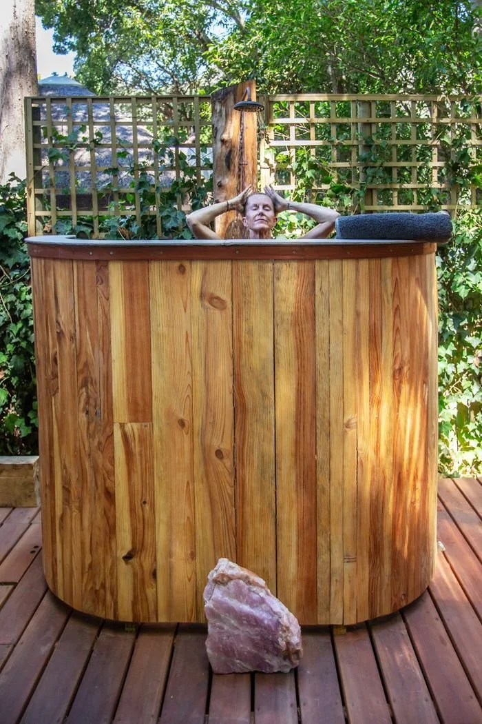 A woman relaxing in a wooden outdoor hot tub on a deck, surrounded by green foliage and fencing, with a large pink rock in front of the tub.