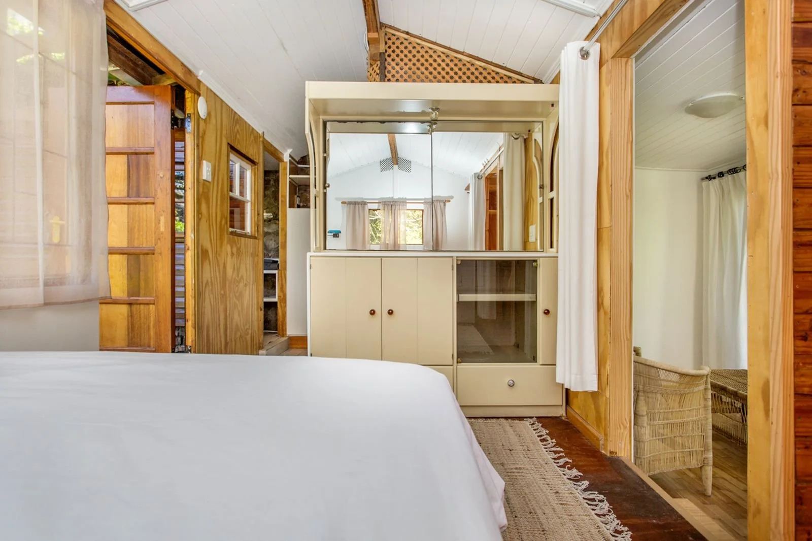 Interior of a cozy bedroom with wooden paneling, a white bed in the foreground, and a small vintage cabinet with a mirror, windows with curtains, and a corner wicker chair.