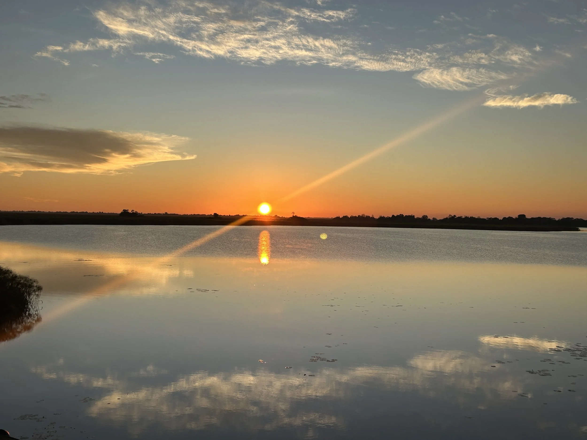 A sunrise over the lagoon in the Delta, casting orange and yellow light. Clouds are scattered across the sky, and the water reflects the sky and sunset colors.
