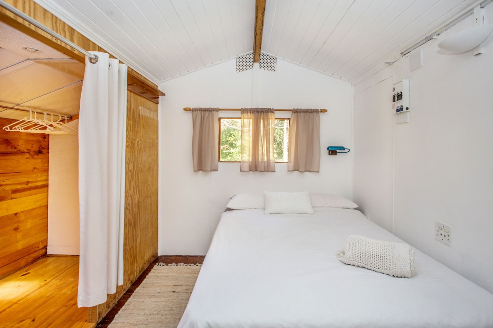 A bedroom with white walls, a bed with white linens, beige curtains on the window, wooden accents, and a closet with a white curtain and hangers.