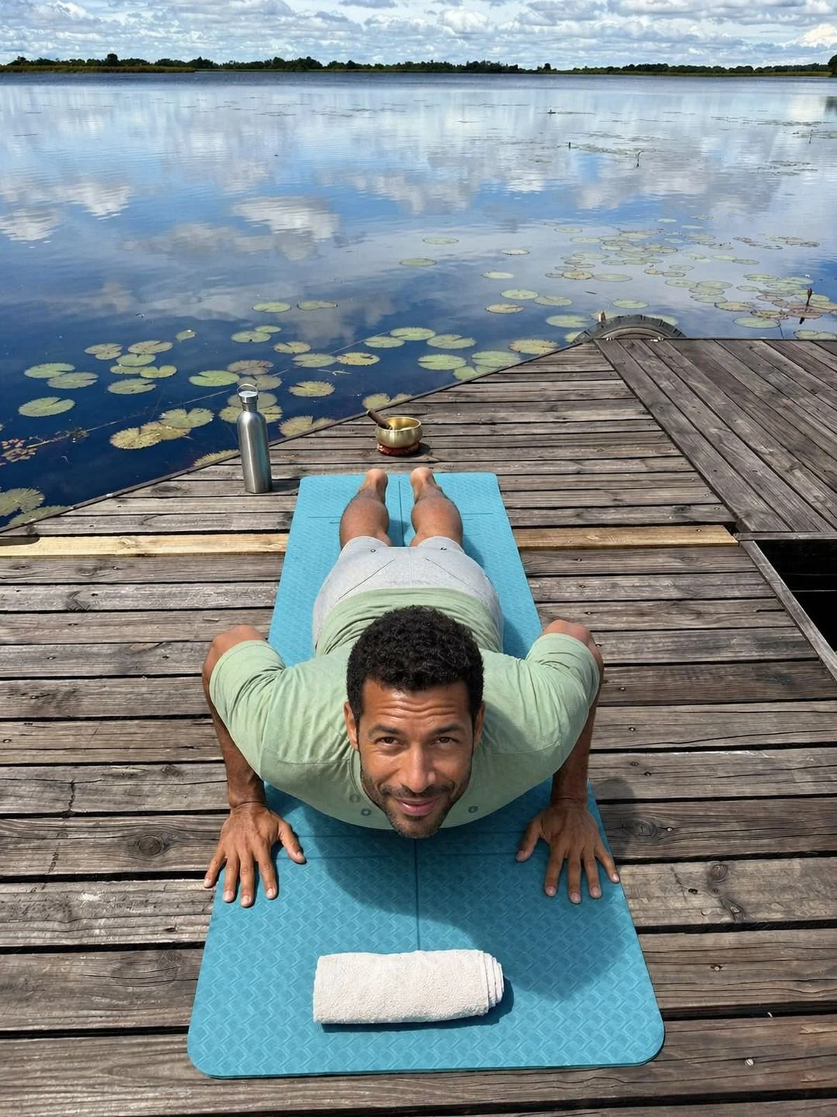 A man practicing yoga on a blue mat on a wooden dock by a lake with lily pads, holding a plank position with a joyful expression, water bottle and singing bowl nearby.