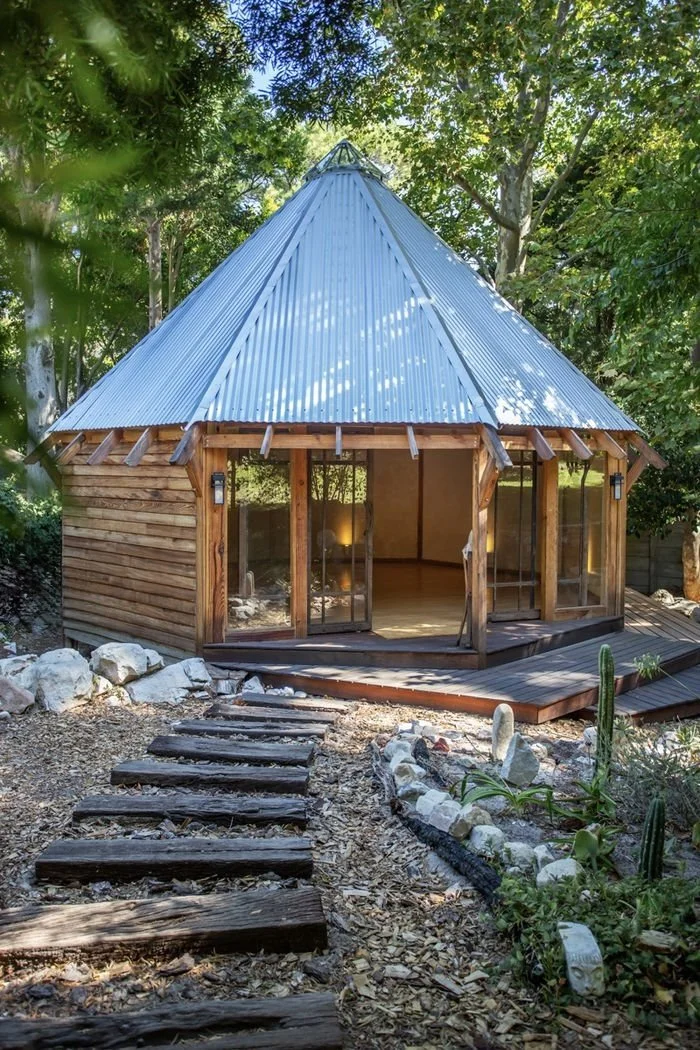 Wooden cabin with a metal roof, surrounded by trees, with a stone and wood pathway leading to a porch with glass doors.