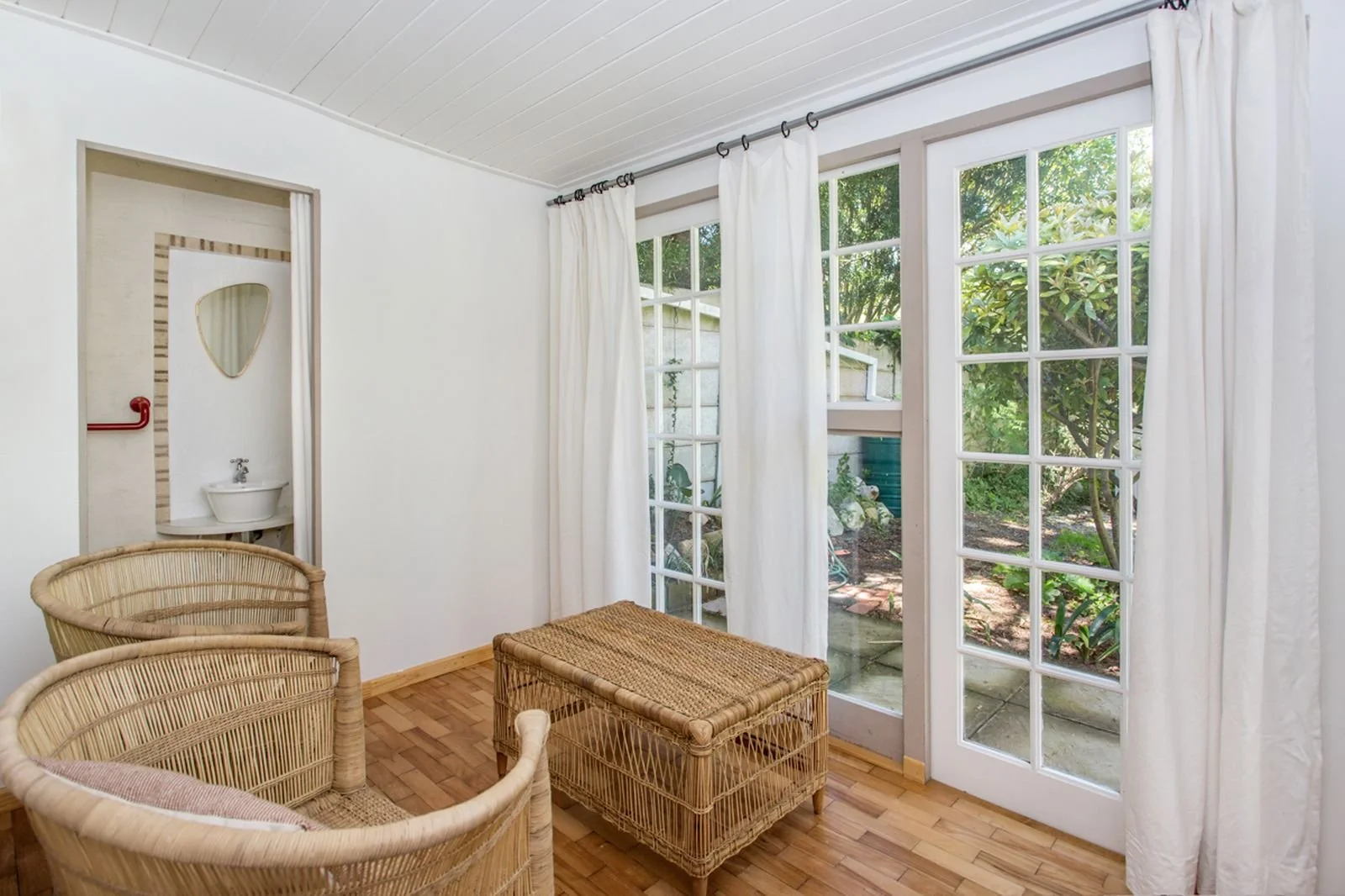 A cozy sunlit room with large window panels and white curtains, a wicker chair, a woven ottoman, and small outdoor garden visible through the windows.