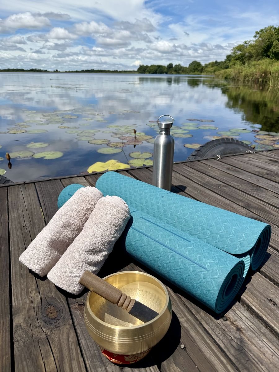 Yoga mats, rolled towels, a singing bowl with a mallet, water bottle, and a towels on a dock overlooking a lake with lily pads and trees in the background, on the largest lagoon in the Delta.