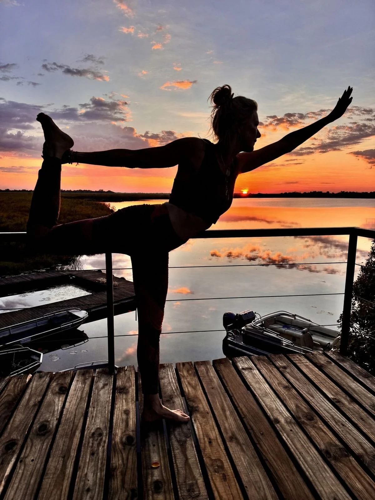 A woman practicing yoga outdoors on a wooden dock at sunset, performing a balancing pose with one leg bent back and held by her hand, near a calm body of water with boats and a scenic sky.