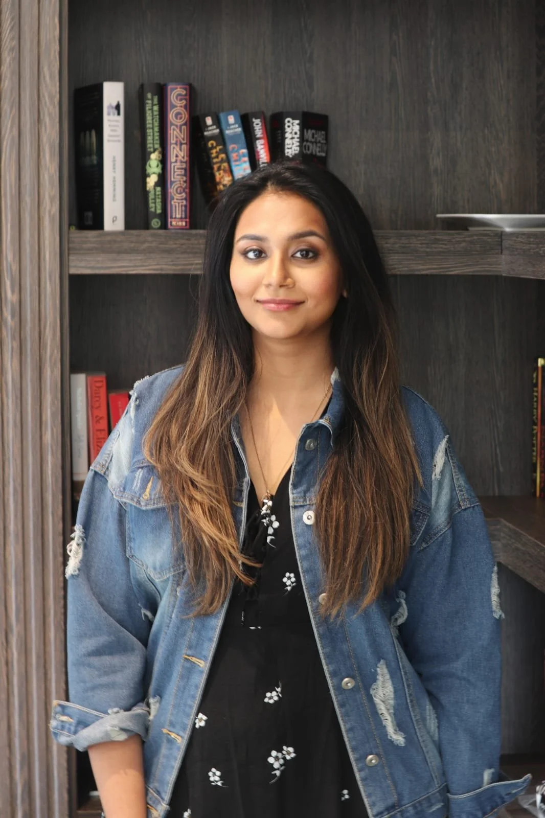Young woman with long, wavy hair wearing a black floral dress and a blue denim jacket standing in front of a dark wood bookshelf filled with books.