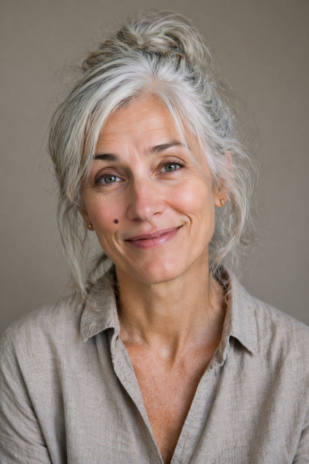 A smiling middle-aged woman with gray and white hair tied up, wearing a beige button-up shirt, looking at the camera.