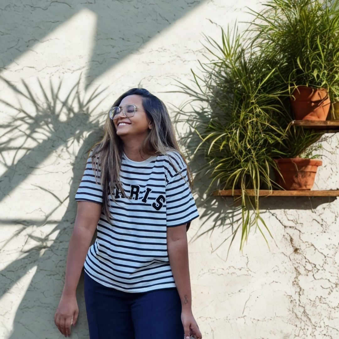 A woman smiling and wearing glasses, standing outdoors against a light-colored textured wall with shadows cast from a large plant. She is dressed in a black and white striped t-shirt with the word 'PARIS' on it and dark pants. There are two shelves on the wall with potted plants.