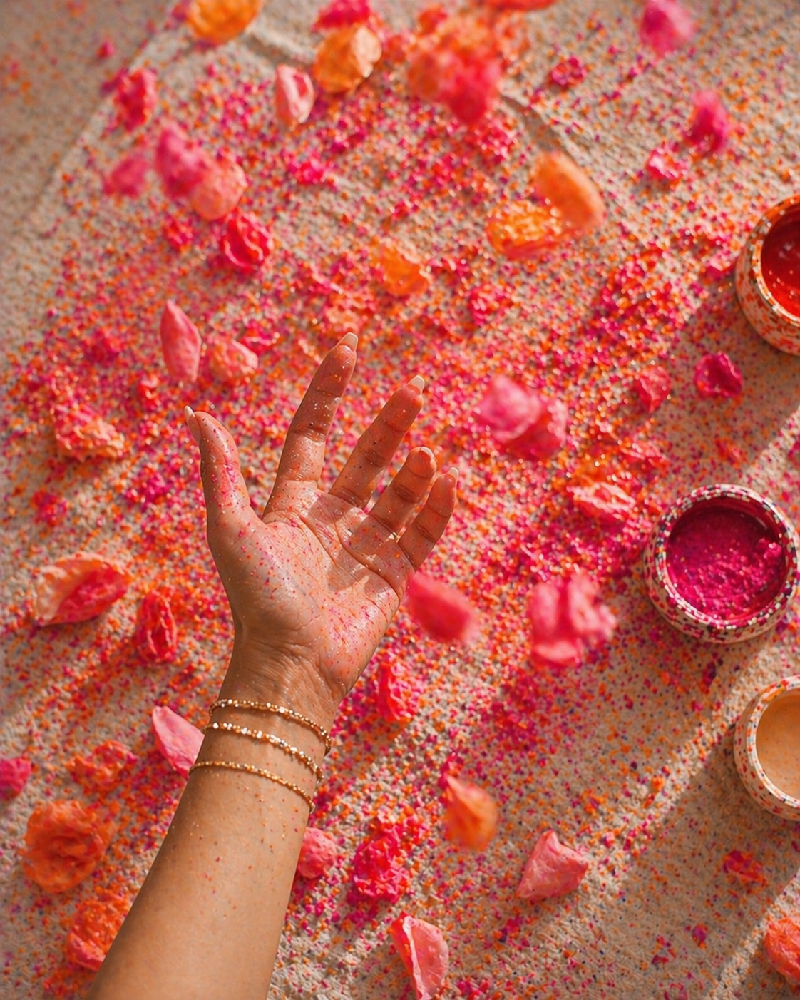 A woman's hand covered in pink and orange sprinkles, with some falling onto a surface filled with pink and orange sprinkles and rose petals, with three small open bowls containing pink, orange, and red powders or pigments to the right.