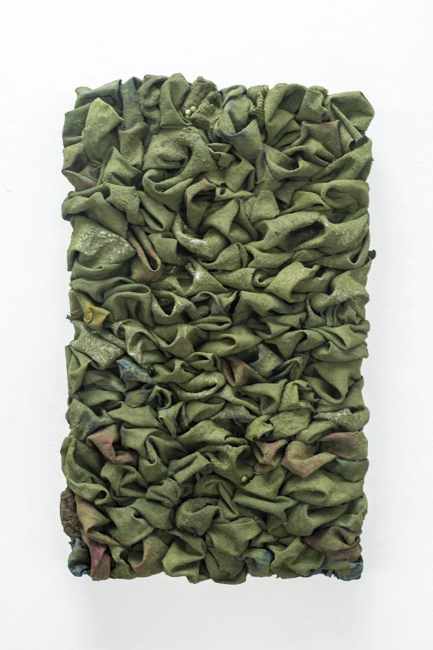 A textured art piece made of crumpled green fabric hung on a white wall. - Emmanuel Aggrey Tieku