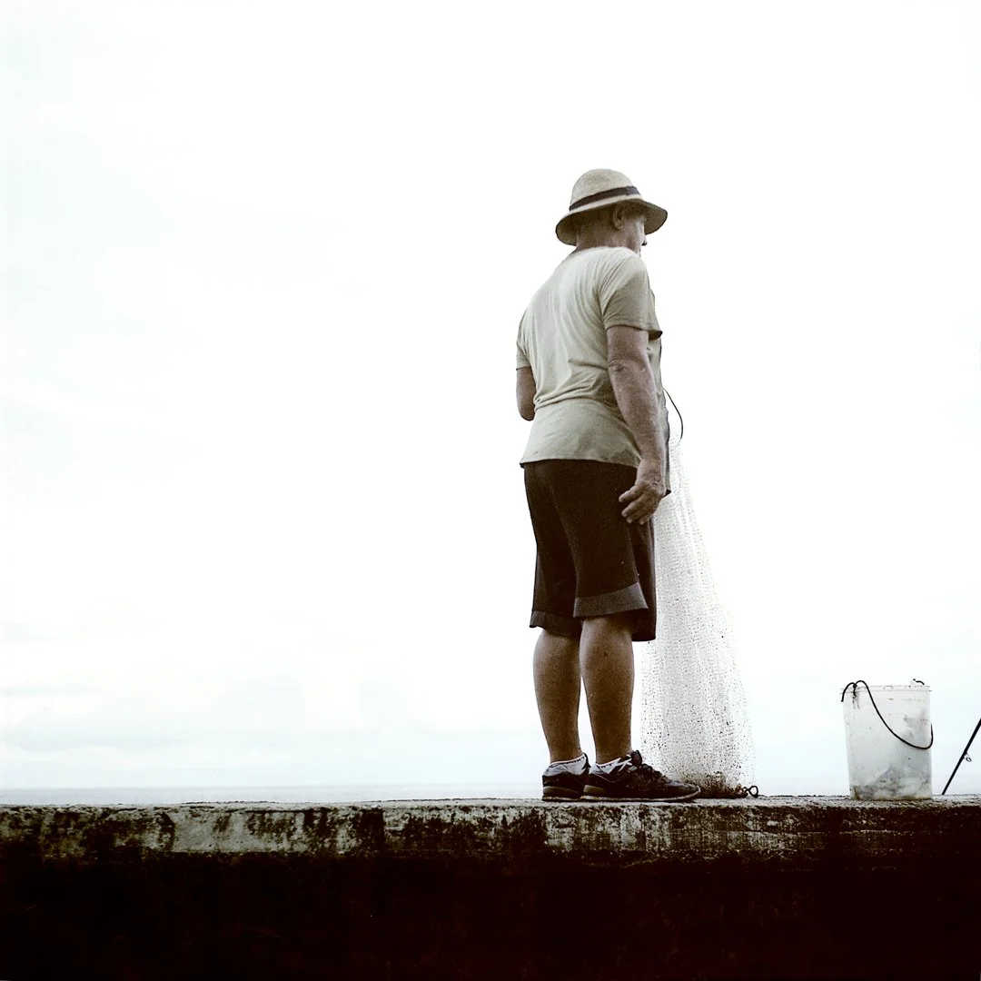 A person standing on a concrete surface near the water, holding a fishing net with a bucket nearby. - Ken Kamara