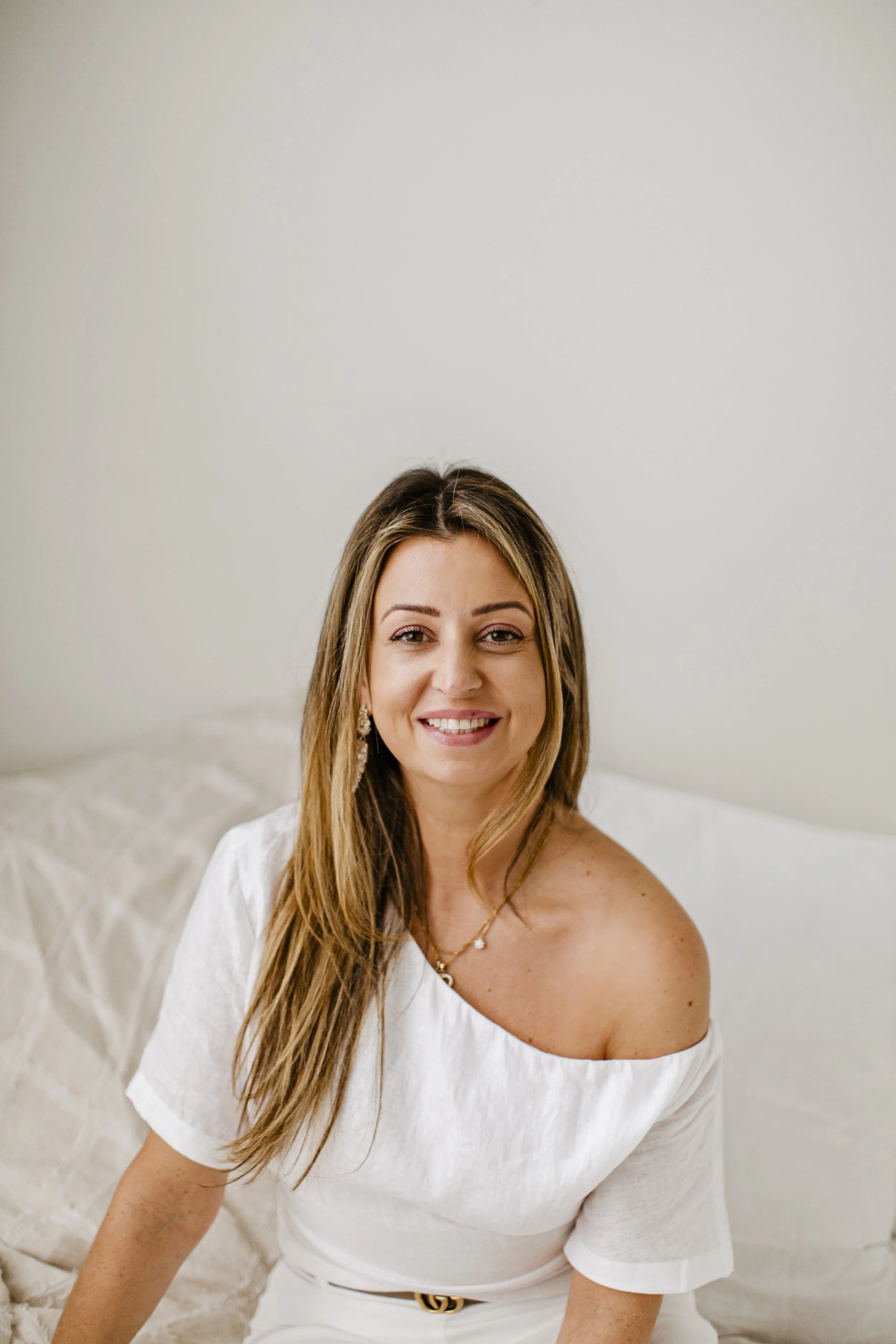 Tash Kolc, cosmetic tattoo artist, smiling in a neutral-toned setting wearing an off-the-shoulder white shirt and gold jewellery.