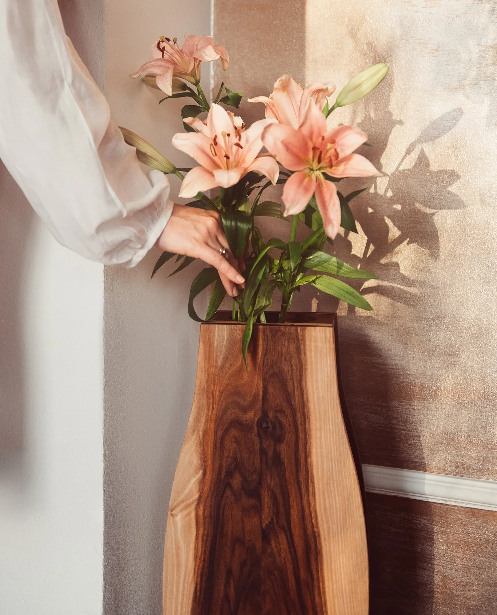 A person's hand holding a bouquet of pink lilies, with some lilies in bloom and one lily bud. The flowers are placed in a wooden vase on the floor, with a textured beige wall in the background and sunlight casting soft shadows.