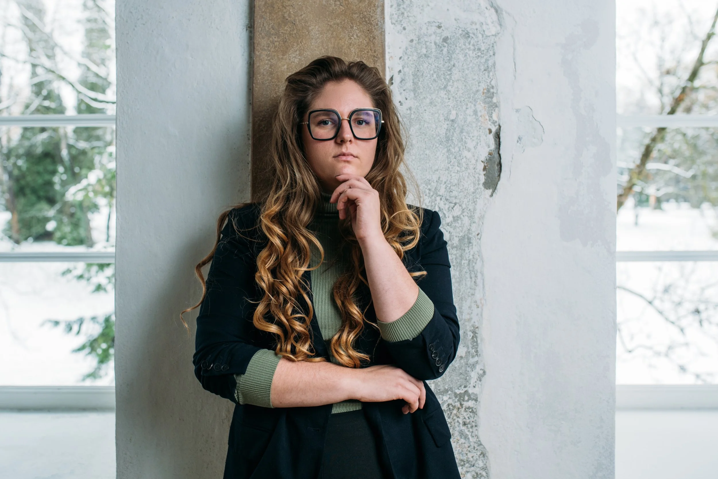 A woman with long, curly hair wearing glasses, a black blazer, and green cuffs, standing inside near a window with trees outside visible. She has one hand on her chin and the other arm crossed in front of her.