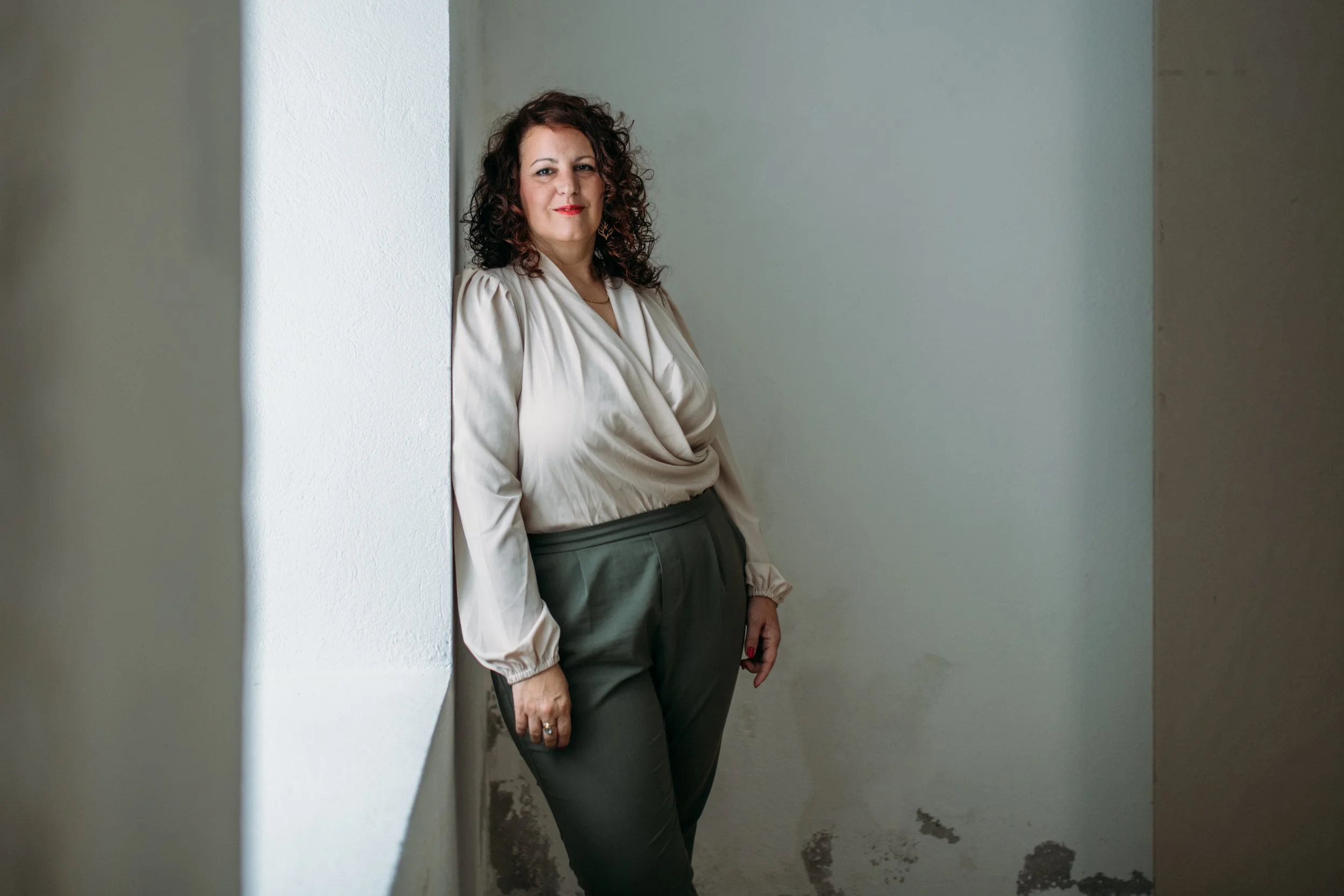 A woman with dark curly hair and red lipstick stands leaning against a white wall in a minimalistic room with plain light-colored walls and a concrete floor.