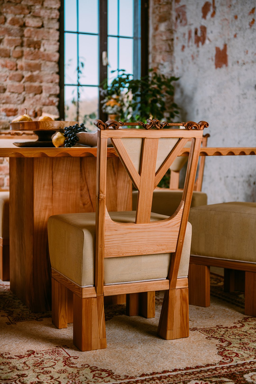 A wooden dining chair with a cushioned seat in front of a dining table. The table has a decorative bowl of fruit, including pears and grapes. The background features a brick wall, a large window with black framing, and indoor plants.