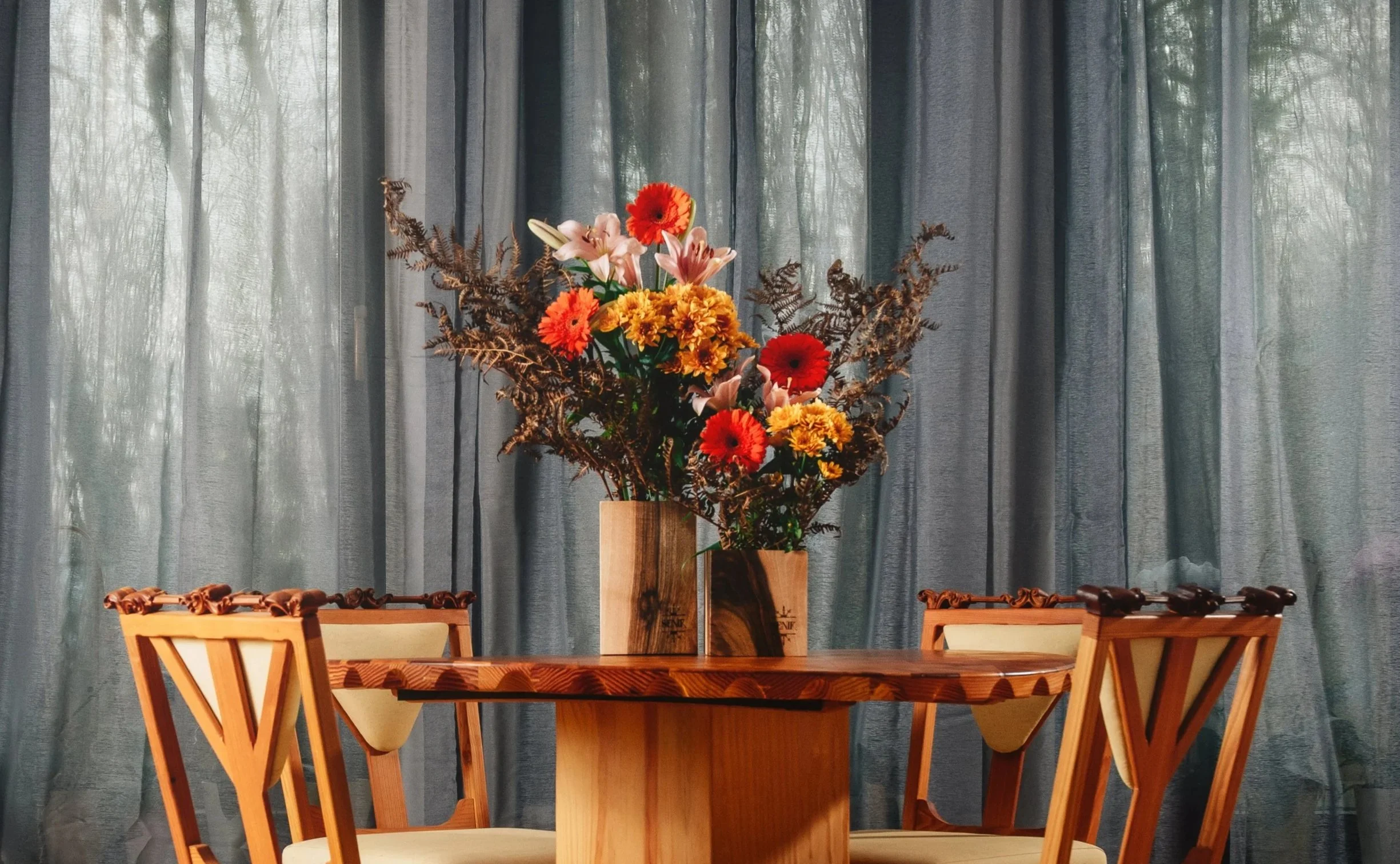 A wooden dining table with a vase of colorful flowers, held in a wooden container, set against a background of gray curtains.